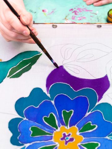 a hand holding a paintbrush and painting a flower on fabric